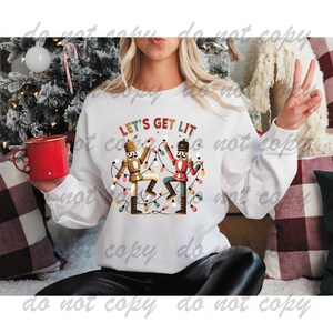May include: White sweatshirt with a Christmas-themed graphic of two nutcracker figures holding string lights and the text "Let's Get Lit".