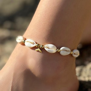 Könnte beinhalten: Fußkettchen mit weißen Muscheln und goldfarbenen Perlen. Das Fußkettchen hat einen kleinen goldfarbenen Anhänger. Der Schmuck wird am Knöchel getragen. Der Hintergrund ist eine verschwommene Szene im Freien.