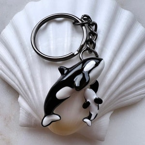May include: A black and white orca whale keychain with a smaller orca attached. The keychain has a silver-colored ring and chain. The orcas are detailed with black and white enamel, and the keychain is set against a white seashell.