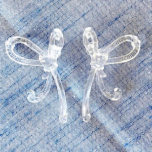 May include: Two clear plastic hair clips shaped like bows. The bows are tied in a knot at the center and have a simple, elegant design.