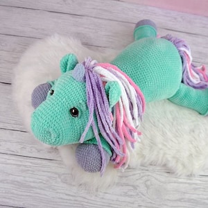 Puede incluir: Un juguete de peluche de unicornio verde tejido a crochet con una larga melena multicolor. El unicornio está acostado sobre una superficie blanca y esponjosa.