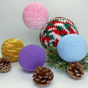 Puede incluir: Cinco adornos de ganchillo en varios colores y patrones. Los adornos son de color rosa, amarillo, morado, azul y rojo y blanco. Los adornos están dispuestos sobre una superficie blanca con ramas de pino y piñas.