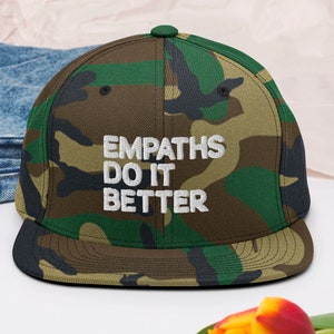 May include: A camouflage baseball cap with the text "Empaths Do It Better" embroidered in white.