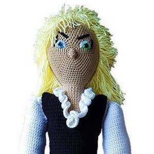 May include: A crocheted doll with long blonde hair, blue eyes, and a white crocheted necklace. The doll is wearing a black and white crocheted dress.