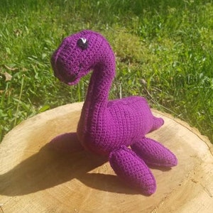May include: A purple crocheted dinosaur toy with black eyes. The dinosaur is sitting on a wooden stump.