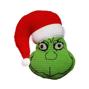 May include: Green crocheted Grinch pillow with a red and white crocheted Santa hat. The Grinch has embroidered eyes, a nose, and a mouth.