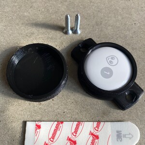 May include: A black plastic round case with a white round disc inside. The disc has a smiley face and the number 7 printed on it. The case has a small loop on the side. Two silver screws and a white adhesive strip with red text are also included.