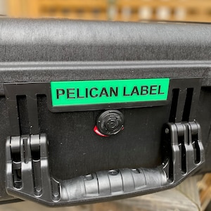 Op de afbeelding: Een zwarte Pelican koffer met een groen label met de tekst "PELICAN LABEL" en een rode sluiting.