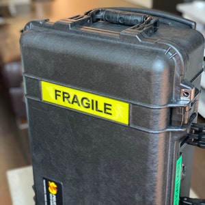 Pode incluir: Uma mala Pelican preta com uma etiqueta amarela "FRAGILE". A mala tem uma superf&iacute;cie texturizada, uma pega preta e um autocolante verde "PELICAN LABEL". Projetada para proteger itens durante o transporte.