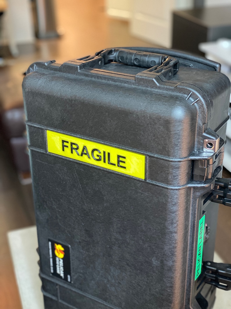 Pode incluir: Uma mala Pelican preta com uma etiqueta amarela "FRAGILE". A mala tem uma superf&iacute;cie texturizada, uma pega preta e um autocolante verde "PELICAN LABEL". Projetada para proteger itens durante o transporte.