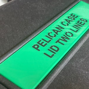 Op de afbeelding: Een zwarte Pelican-koffer met een groen label waarop "PELICAN CASE LID TWO LINES" staat.