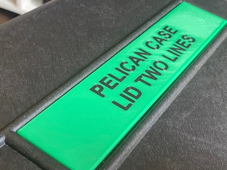 Op de afbeelding: Een zwarte Pelican-koffer met een groen label waarop "PELICAN CASE LID TWO LINES" staat.