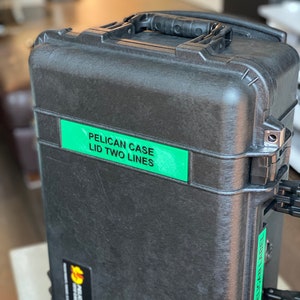 Op de afbeelding: Een zwarte Pelican-koffer met een groen label met de tekst "PELICAN CASE LID TWO LINES".