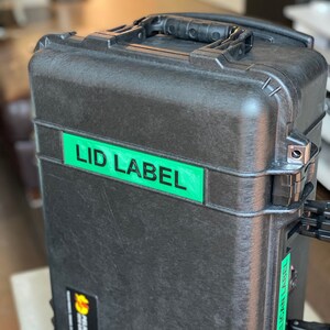 Pode incluir: Uma mala preta e resistente com uma pega e etiquetas verdes que dizem "LID LABEL" e "PELICAN LABEL". A mala tem uma superf&iacute;cie texturizada e fechos met&aacute;licos. Um pequeno autocolante com o log&oacute;tipo Pelican &eacute; vis&iacute;vel.