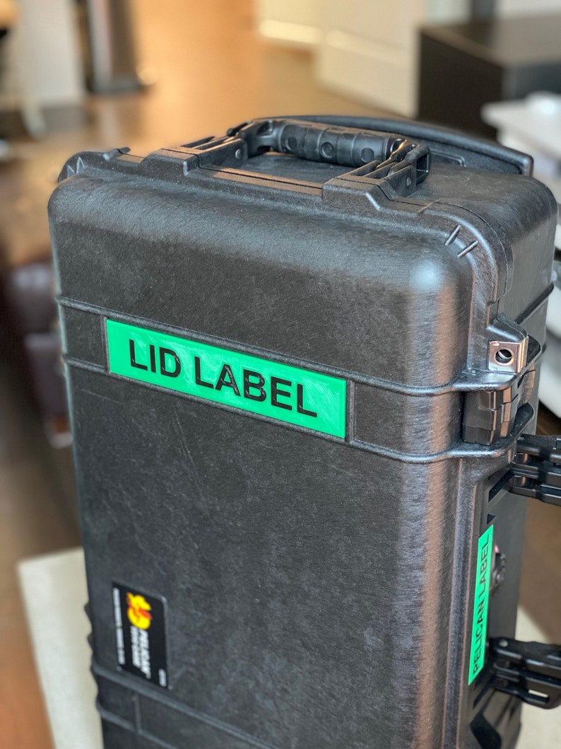 Pode incluir: Uma mala preta e resistente com uma pega e etiquetas verdes que dizem "LID LABEL" e "PELICAN LABEL". A mala tem uma superf&iacute;cie texturizada e fechos met&aacute;licos. Um pequeno autocolante com o log&oacute;tipo Pelican &eacute; vis&iacute;vel.