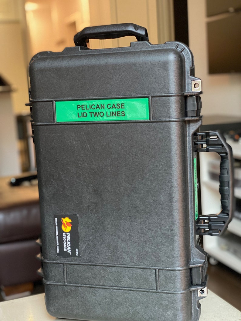 Op de afbeelding: Een zwarte Pelican-koffer met een groen label met de tekst "Pelican Case Lid Two Lines".