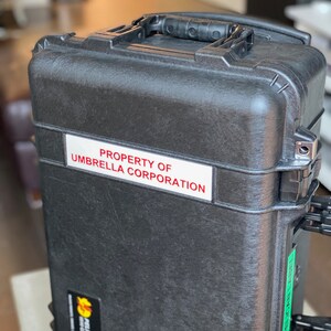 Op de afbeelding: Een zwarte harde koffer met een handvat en een witte sticker met de tekst "PROPERTY OF UMBRELLA CORPORATION".