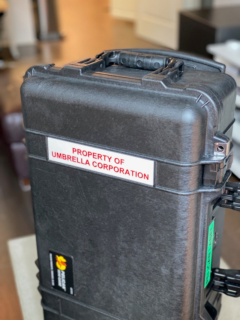 Op de afbeelding: Een zwarte harde koffer met een handvat en een witte sticker met de tekst "PROPERTY OF UMBRELLA CORPORATION".
