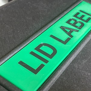 Pode incluir: Um objeto retangular preto com uma etiqueta verde que diz "LID LABEL" em texto preto. A etiqueta est&aacute; centrada e tem uma apar&ecirc;ncia impressa em 3D em relevo. O fundo &eacute; uma superf&iacute;cie escura texturizada.