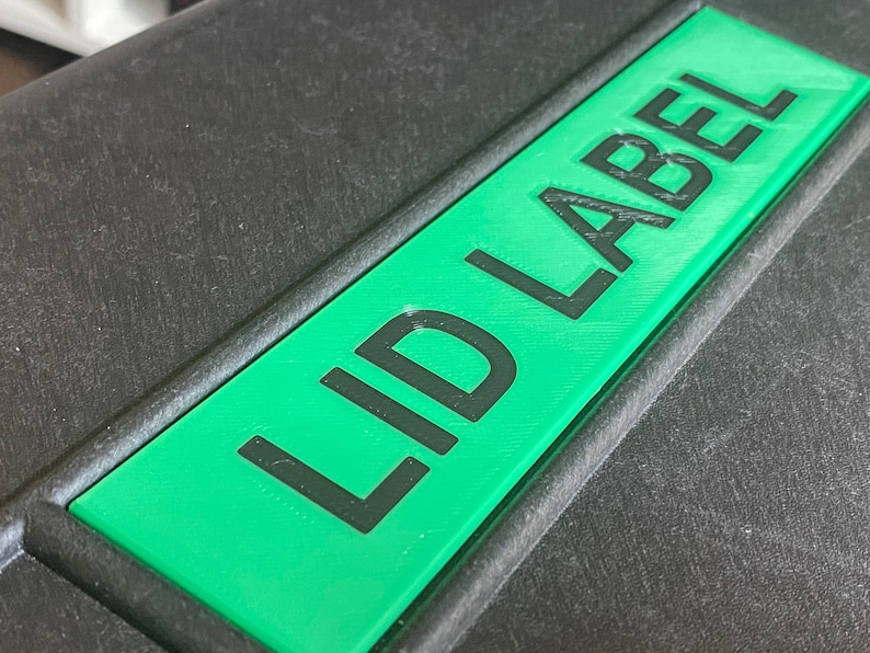Pode incluir: Um objeto retangular preto com uma etiqueta verde que diz "LID LABEL" em texto preto. A etiqueta est&aacute; centrada e tem uma apar&ecirc;ncia impressa em 3D em relevo. O fundo &eacute; uma superf&iacute;cie escura texturizada.
