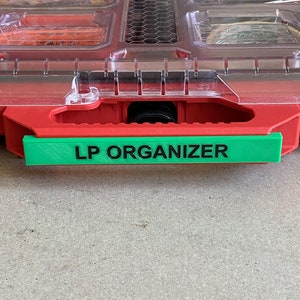 Könnte beinhalten: Rote und transparente Kunststoff-Aufbewahrungsbox mit einem grünen Etikett mit der Aufschrift "LP ORGANIZER".