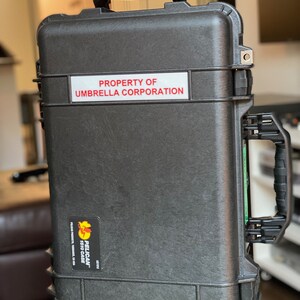 Op de afbeelding: Een zwarte Pelican 1610 koffer met een sticker waarop staat "PROPERTY OF UMBRELLA CORPORATION".