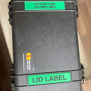 Pode incluir: Uma mala preta Pelican 1510 com etiquetas verdes que dizem "PELICAN CASE LID TWO LINES" e "LID LABEL". A mala tem um adesivo Pelican Products. A mala retangular tem um fecho de encaixe.