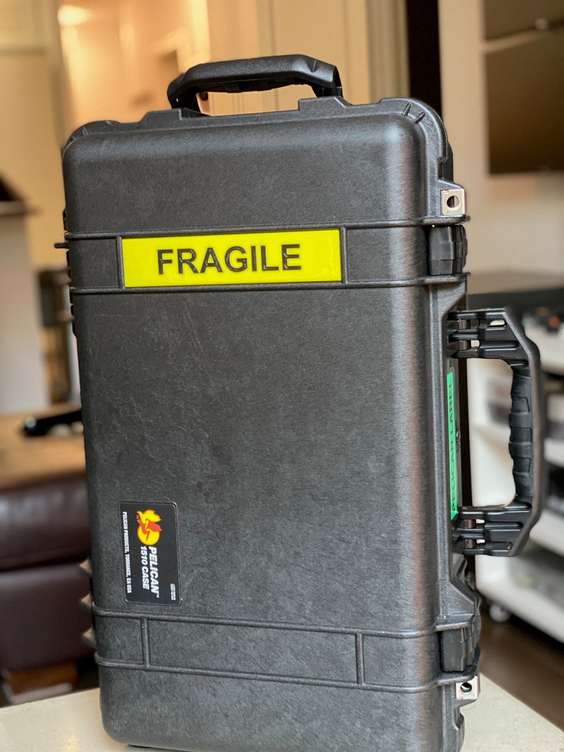 Pode incluir: Uma mala preta Pelican 1510 com um autocolante amarelo "FRAGILE". A mala tem uma pega na parte superior e uma pega lateral. Um autocolante Pelican Products &eacute; vis&iacute;vel na frente. A mala foi concebida para proteger equipamentos sens&iacute;veis.