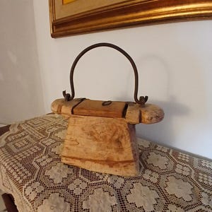 Könnte beinhalten: Ein rustikaler Holzeimer mit einem Metallgriff. Der Eimer besteht aus hellfarbenem Holz und hat einen gebogenen Metallgriff, der oben befestigt ist. Der Eimer steht auf einem Tisch, der mit einer weißen Spitzentischdecke bedeckt ist. Im Hintergrund hängt ein gerahmtes Gemälde.