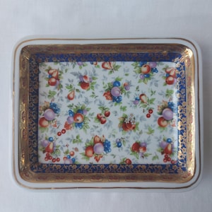 Peut inclure: Plateau rectangulaire en céramique avec un fond blanc et un motif de fruits colorés. Le plateau a une bordure bleue avec des accents dorés et un bord doré. Le motif de fruits comprend des pommes, des poires, des prunes et des baies.