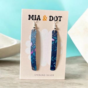 May include: A pair of long, thin, rectangular dangle earrings featuring a dark blue background with a repeating pattern of small leaves in shades of light blue, pink, and white. The earrings are attached to a beige display card labelled MIA & DOT.