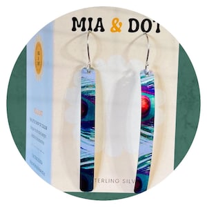 May include: A pair of long, rectangular earrings with a peacock feather design in shades of blue, green, and red. The earrings are suspended from silver hooks. The packaging reads "MIA & DOT" and "STERLING SILVER".