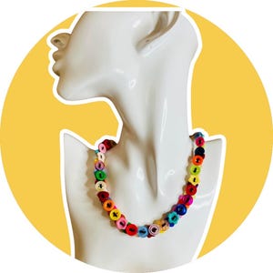 Puede incluir: Un collar de botones coloridos exhibido en un maniquí blanco. El collar presenta una variedad de botones de colores brillantes, incluyendo rojo, azul, verde, amarillo y rosa. Los botones están ensartados para crear un accesorio único y llamativo.