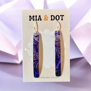 Pode incluir: Um par de brincos pendentes retangulares, longos e finos, com um padrão abstrato em roxo escuro, preto e cinzento. Os brincos estão pendurados em ganchos prateados e exibidos num cartão bege com a etiqueta MIA & DOT e STERLING SILVER.