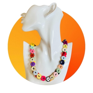 Op de afbeelding: Een kleurrijke knopenketting tentoongesteld op een witte mannequin buste. De ketting heeft verschillende knopen in verschillende kleuren en patronen, waaronder rood, roze, geel en blauw. De achtergrond is een heldere oranje cirkel.