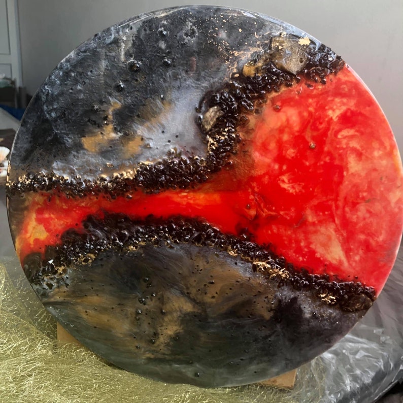 May include: A round, black, white, gold, and red resin art piece with a swirling, abstract design. The piece has a glossy finish and is reminiscent of a galaxy or a fiery explosion.