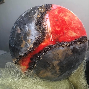 May include: A round, abstract art piece with a black and red epoxy resin design. The resin has a glossy finish and features gold accents.