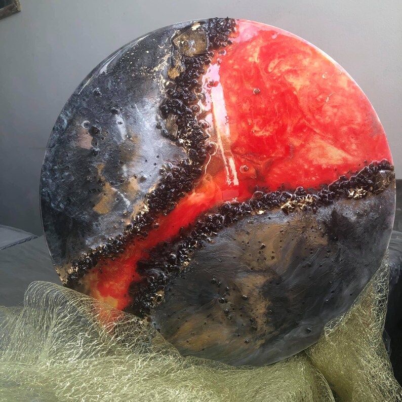 May include: A round, abstract art piece with a black and red epoxy resin design. The resin has a glossy finish and features gold accents.