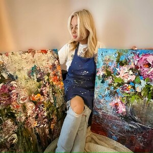 May include: Two abstract floral paintings with a textured, impasto style. The paintings are in shades of blue, green, pink, and white. The artist is sitting in front of the paintings, wearing a blue apron and jeans.
