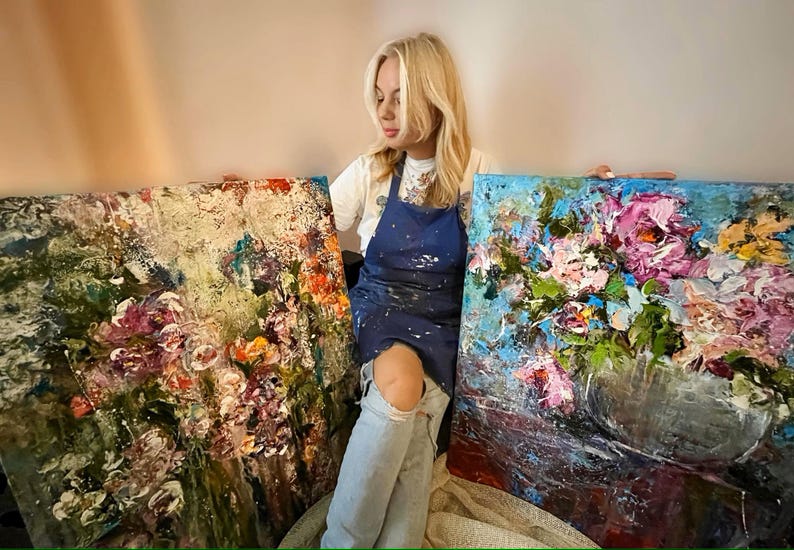 May include: Two abstract floral paintings with a textured, impasto style. The paintings are in shades of blue, green, pink, and white. The artist is sitting in front of the paintings, wearing a blue apron and jeans.