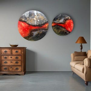 May include: Two round abstract paintings with a red, black, and gold color scheme. The paintings are hanging on a gray wall above a wooden dresser and a brown couch.
