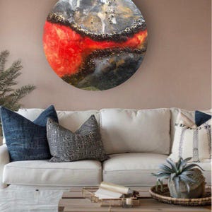 May include: A round, abstract painting with a red, black, and gold color scheme. The painting is mounted on a wall above a white couch with pillows.