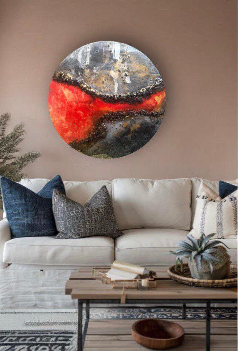 May include: A round, abstract painting with a red, black, and gold color scheme. The painting is mounted on a wall above a white couch with pillows.