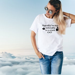 Puede incluir: Camiseta blanca con texto negro que dice "Físicamente estoy aquí, Emocionalmente estoy en un libro". El texto está acompañado por una taza de café, un corazón y una pila de libros.