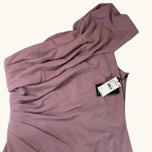 Puede incluir: Un vestido de un solo hombro en color rosa empolvado con detalles de tela drapeada. El vestido presenta un corpi&ntilde;o ajustado y un escote asim&eacute;trico fluido. Una etiqueta negra est&aacute; adherida al vestido.