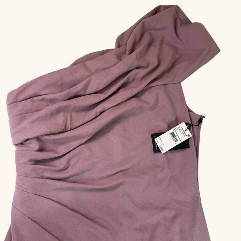 Puede incluir: Un vestido de un solo hombro en color rosa empolvado con detalles de tela drapeada. El vestido presenta un corpi&ntilde;o ajustado y un escote asim&eacute;trico fluido. Una etiqueta negra est&aacute; adherida al vestido.