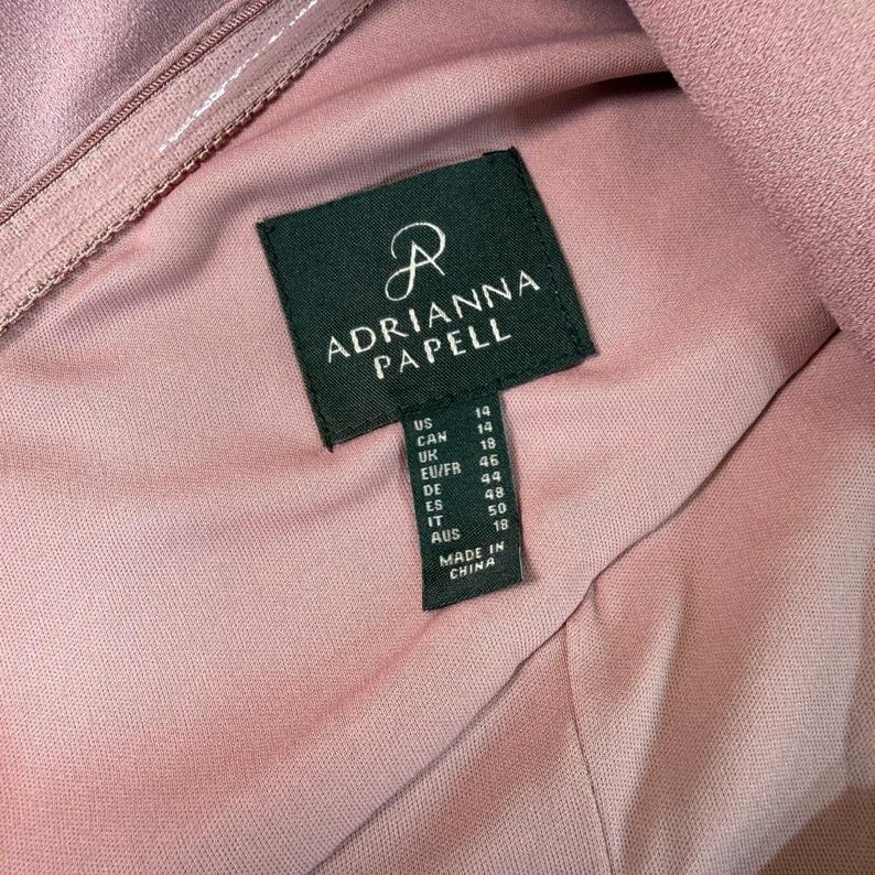 Puede incluir: Primer plano de un vestido color rosa empolvado con una etiqueta de Adrianna Papell. La etiqueta verde oscuro muestra tallas para US, CAN, UK, EU/FR, DE, ES, IT y AUS. El vestido est&aacute; hecho en China.