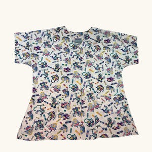 May include: White scrub top featuring a vibrant pattern of cartoon figures, skateboards, and other playful designs. The top has short sleeves, a V-neck, and two front pockets. The design incorporates shades of blue, green, yellow, and pink.