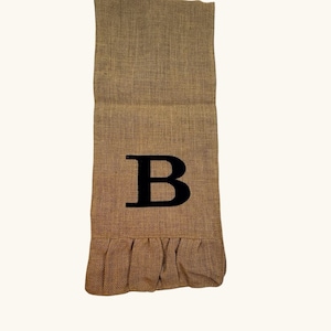 Puede incluir: Camino de mesa de arpillera marrón con una letra "B" negra en el centro. El borde inferior tiene un detalle con volantes. El camino de mesa está hecho de tela texturizada y es adecuado para la decoración del hogar.