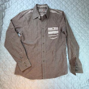 May include: A brown long-sleeved shirt with white embroidered detailing on the shoulders, collar, and pocket. The shirt has a button-down front, cuffs, and a chest pocket. The fabric has a textured appearance. The shirt is a medium size.
